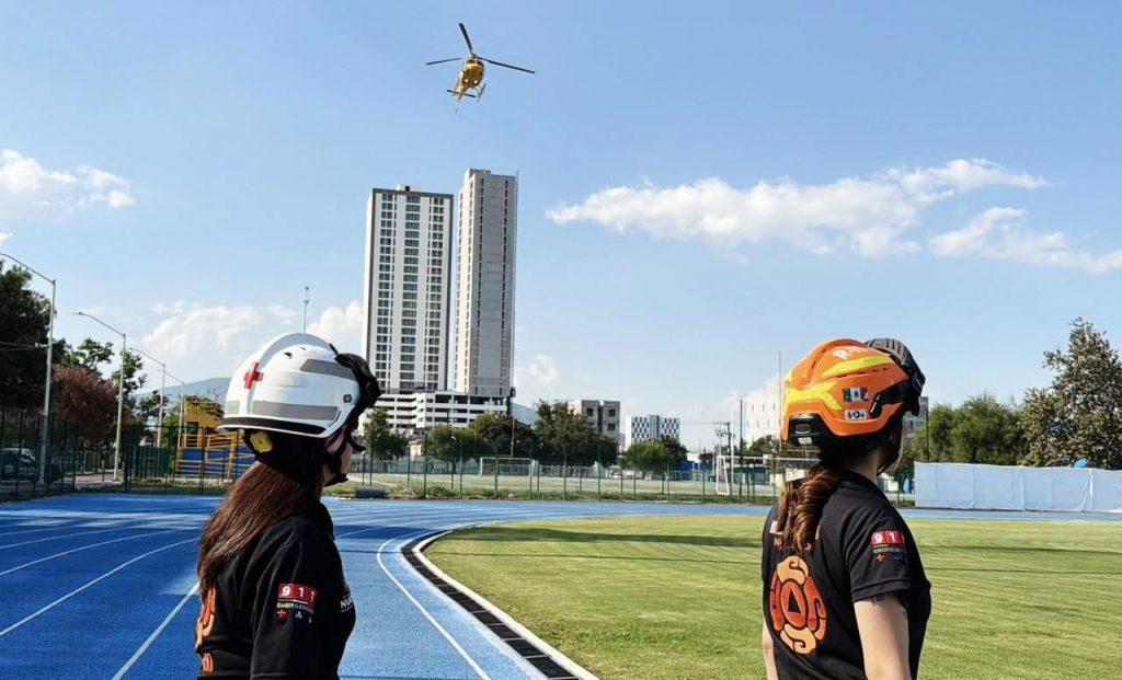 Two women in helmeted gear stand on a blue running track watching a yellow helicopter hover in a clear sky above a cityscape   Informe Regio