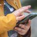 Person in a mustard yellow jacket taps the screen of a smartphone outdoors showing hands and device   Informe Regio
