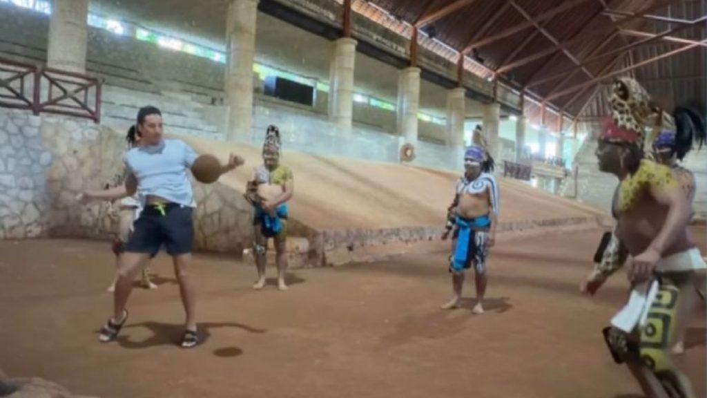 Man in a light blue shirt and dark shorts juggles two brown balls in a large indoor arena watched by Indigenous dancers in feathered headdresses and regalia   Informe Regio