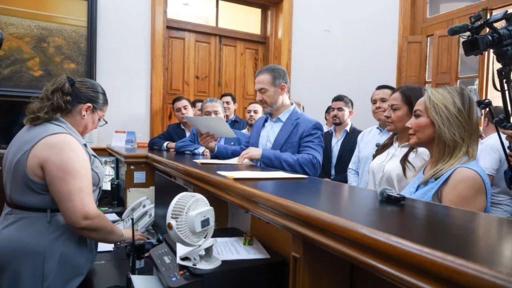 A man in a blue suit reads documents at a dark desk while a group of officials and a camera crew watch in a formal room   Informe Regio