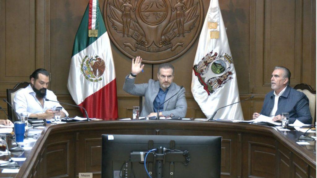 Central man raises his hand during a formal meeting at a curved wooden table with two flags behind him Mexico and another emblem   Informe Regio