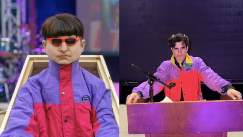 Stage scene with two performers a life size doll like figure in a purple and red jacket with red sunglasses on the left and a man in a matching jacket at a purple podium with a microphone on the right   Informe Regio