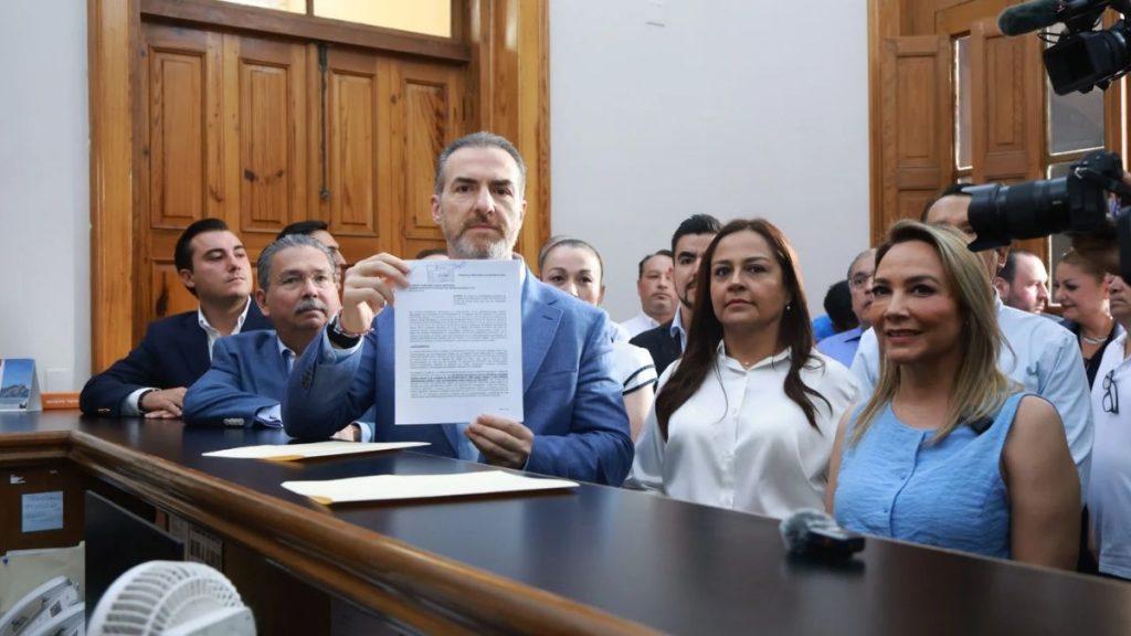 Man in a blue blazer holds up a document in a wood paneled meeting room flanked by officials and reporters with cameras behind him   Informe Regio