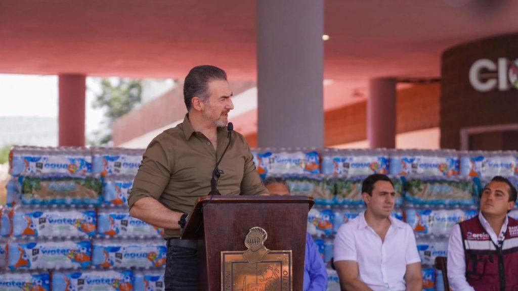 Man in an olive shirt speaks from a wooden podium with a microphone at an outdoor event stacked water packs visible in the background   Informe Regio