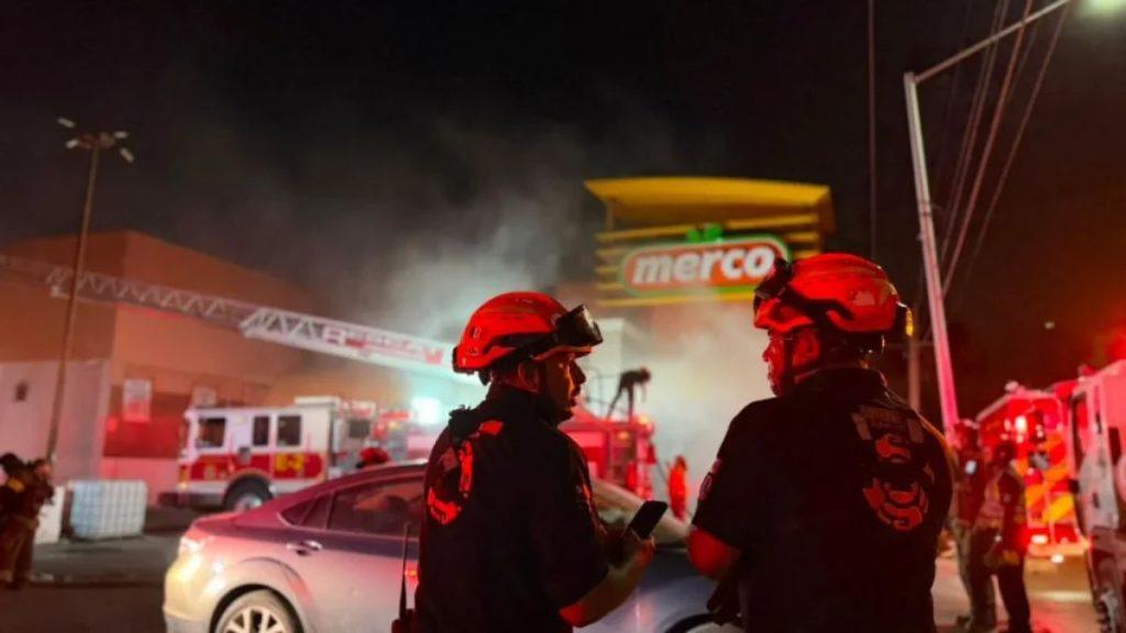 Fuerte-incendio-monterrey-mercado-merco - Informe Regio Firefighters in gear converse near a car at a nighttime fire scene with smoke and emergency lights in the background Informe Regio