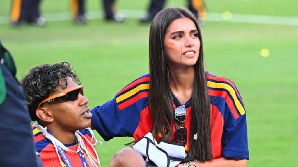 Two young sports fans seated on a grass field in red blue and yellow jerseys woman has face paint and sunglasses tucked in her shirt while the man wears an orange visor and sunglasses nearby   Informe Regio