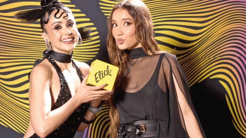 Two women pose at a bright event one in a sequined black dress holding a yellow box that says Eliot   Informe Regio