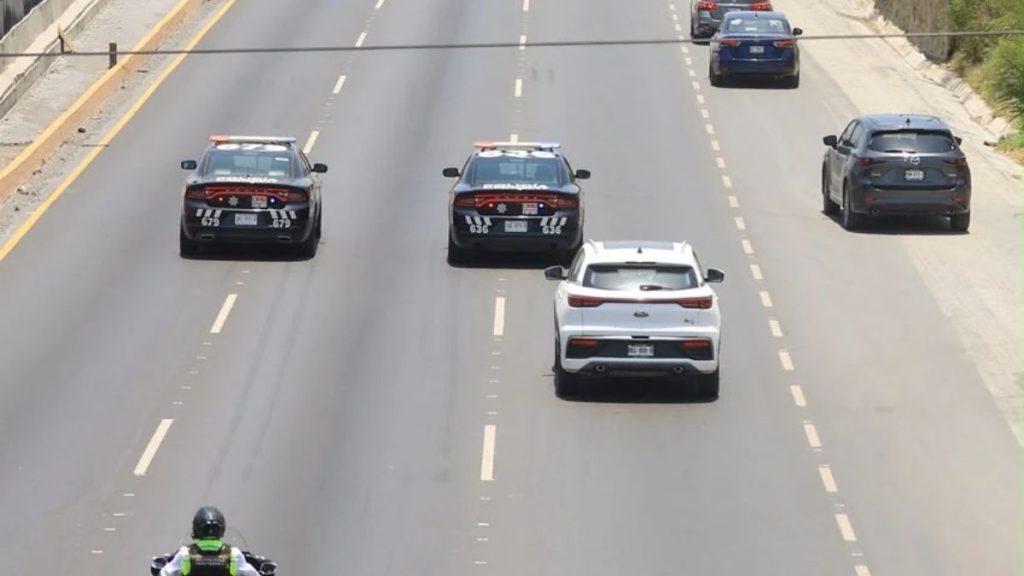 Multa-exceso-velocidad-monterrey - Informe Regio On a multi lane highway two police cars with flashing lights tail a white SUV in the middle lane with other vehicles ahead and a motorcyclist in the bottom left foreground Informe Regio
