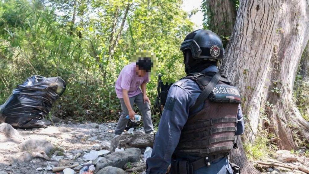 Police officer in tactical gear faces a man collecting a bottle in a litter strewn rocky wooded area   Informe Regio
