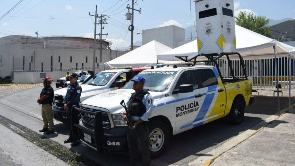 Policía-de-monterrey-joven-15-años - Informe Regio Police officers stand beside a white and blue Monterrey police pickup with a tall armored device on the bed in a street with white tents nearby Informe Regio