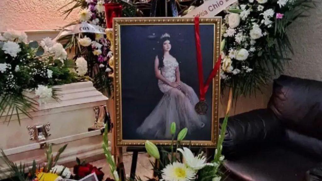 Framed portrait of a woman in a white gown and crown on an easel surrounded by funeral flowers and a hanging medal ribbon on display at a memorial   Informe Regio