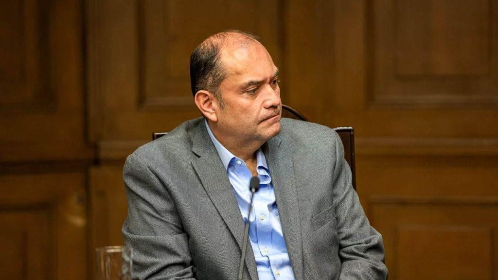 Acuerdo-juridico-gobierno-monterrey-colegio-abogados - Informe Regio Man in a gray suit seated at a table with a microphone in front wood paneled backdrop looking to his left during a formal hearing or meeting Informe Regio