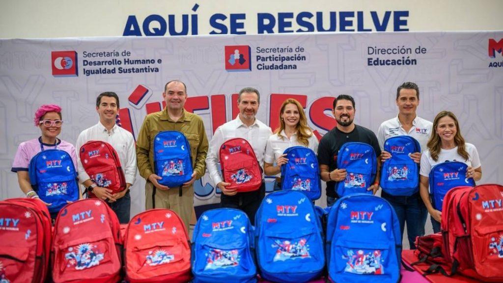 Adrian-de-la-garza-utiles-gratis-monterrey - Informe Regio Eight people stand in a row each holding blue or red backpacks in front of a backdrop with official logos and the words AQUÍ SE RESUELVE visible Informe Regio