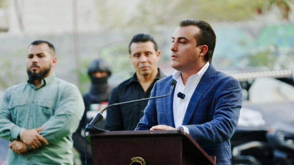 Man in a blue blazer speaks at a podium outdoors with two men standing behind him one in a green shirt the other in black   Informe Regio