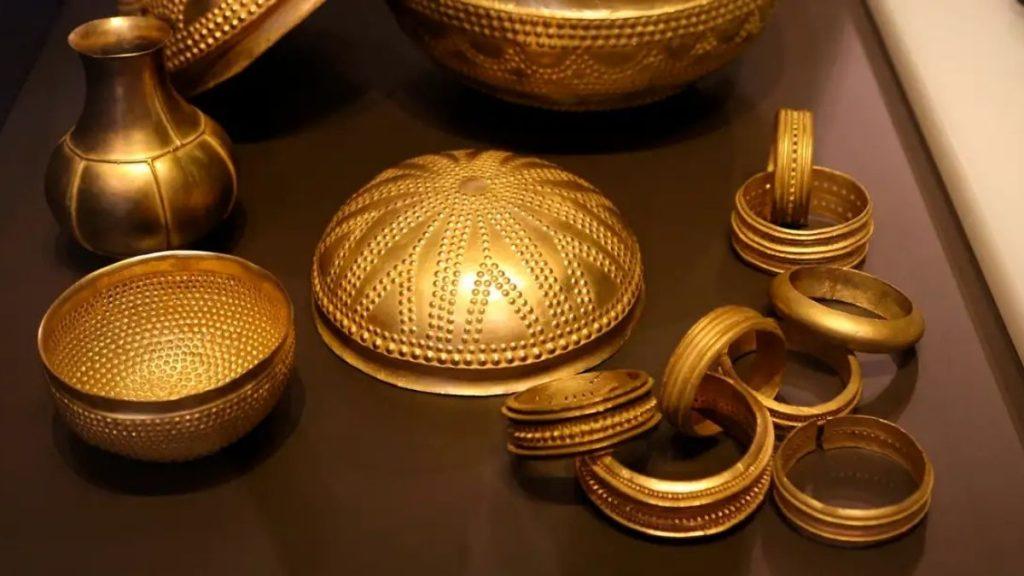 Assorted gold jewelry on a dark surface bangles a domed decorative lid and a small vase like piece in the background   Informe Regio