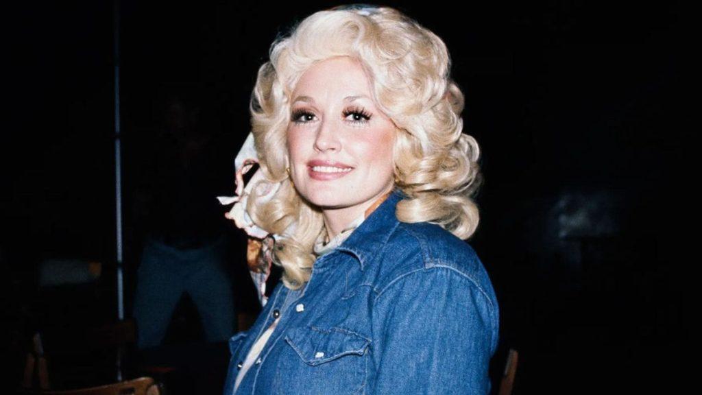Dolly-parton-fallece-a-los-80-años - Informe Regio Smiling woman with voluminous platinum blonde curls wearing a denim jacket against a dark background Informe Regio