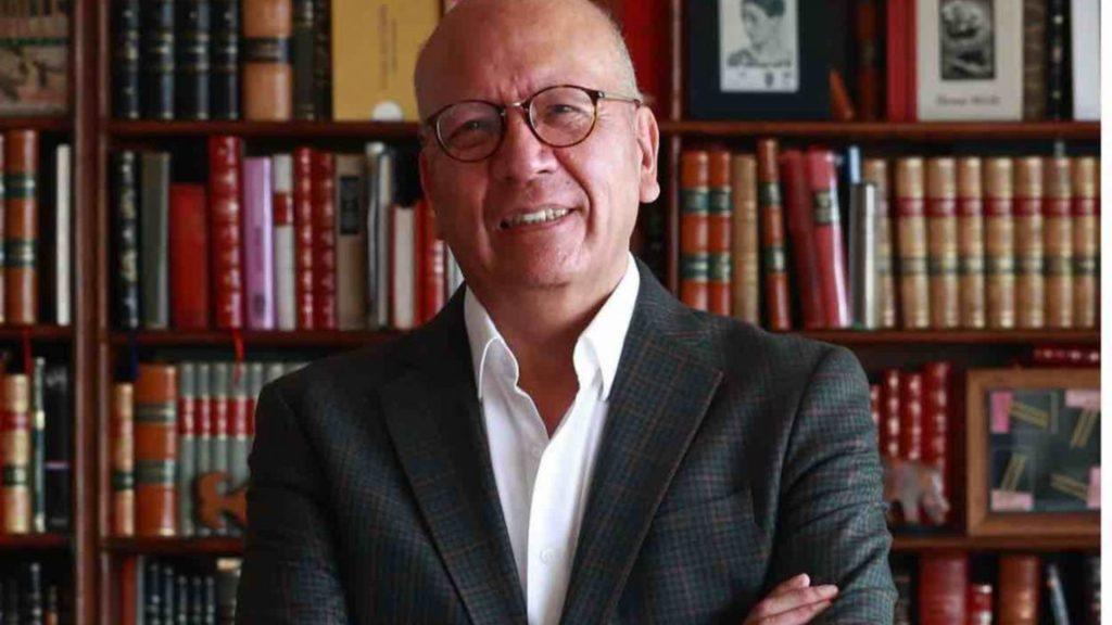 Smiling older man with glasses in a blazer standing in front of a wooden bookshelf filled with books   Informe Regio