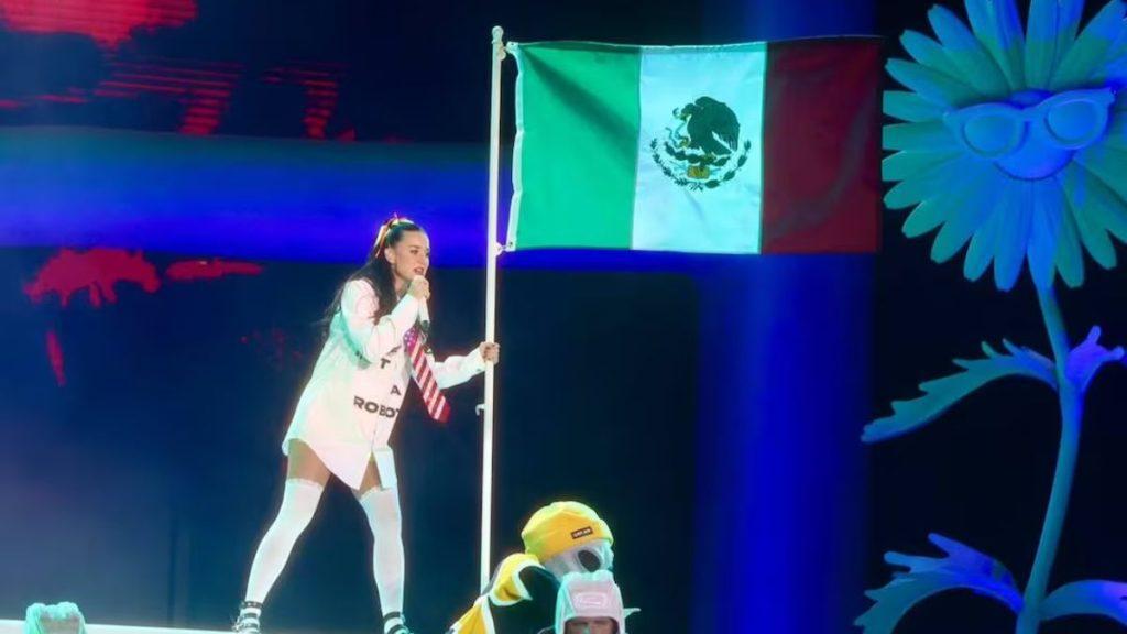 Female performer on a brightly lit stage holds a Mexican flag on a pole wearing a white outfit and knee high socks with colorful stage decor in the background   Informe Regio