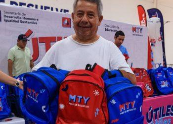 Man smiles while handing out blue and red MTY backpacks at a charity event table full of backpacks behind him   Informe Regio