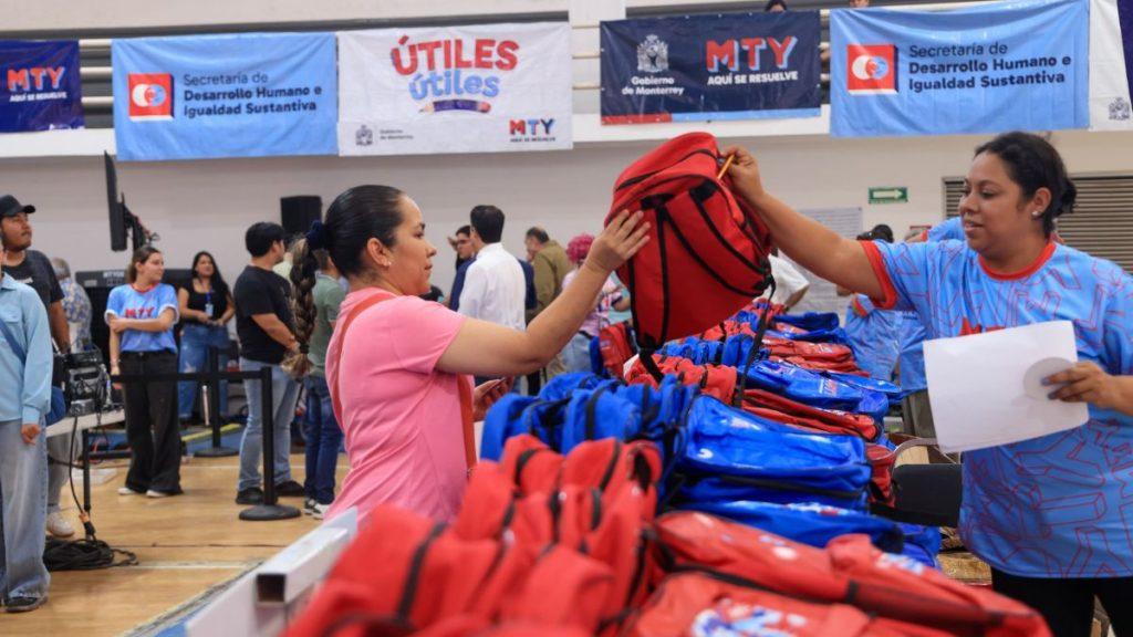 Utiles-utiles-monterrey - Informe Regio Woman hands a red backpack to a participant at a backpack distribution event stacks of red and blue bags and banners in the background Informe Regio