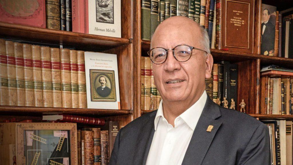 Older man in a suit and round glasses smiling standing in a library with wooden shelves full of old books   Informe Regio