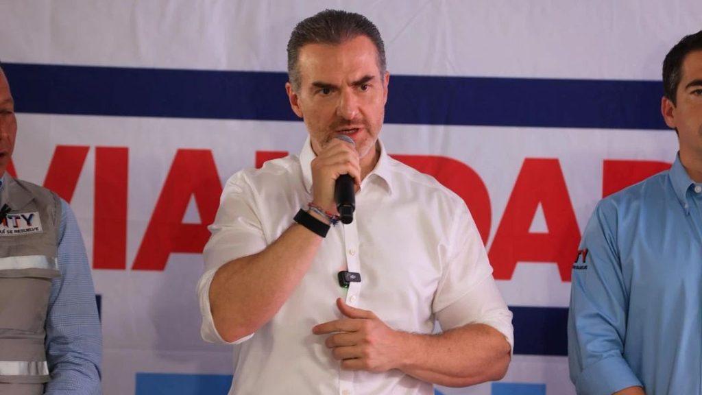 Man in a white shirt speaks into a handheld microphone on a stage flanked by two men in blue and gray outfits in front of a banner behind him   Informe Regio