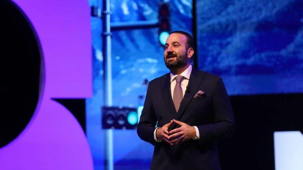 Male speaker in a dark suit and tie delivering a talk on a stage with purple lighting and a lapel mic   Informe Regio