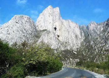 Curved mountain road at the base of towering white limestone cliffs under a blue sky, with green bushes along the roadside.