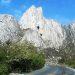 Curved mountain road at the base of towering white limestone cliffs under a blue sky with green bushes along the roadside   Informe Regio