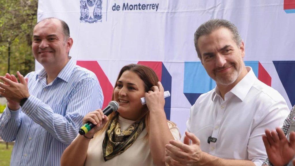 Three people on a stage at an outdoor event a woman speaks into a microphone while two men applaud nearby   Informe Regio