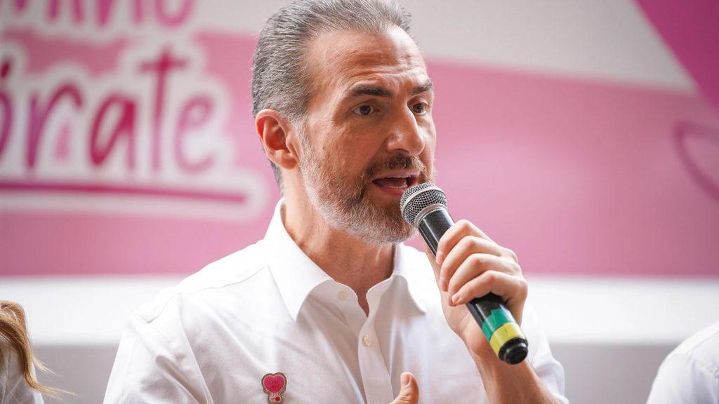 Man with a gray beard in a white shirt speaks into a handheld microphone at a pink themed event   Informe Regio