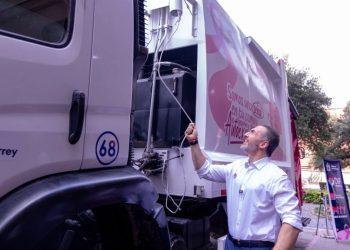 Man in a white shirt stands beside a large white truck with pink branding, reaching up to connect a cable to the vehicle's equipment outside a building complex at a daytime event.