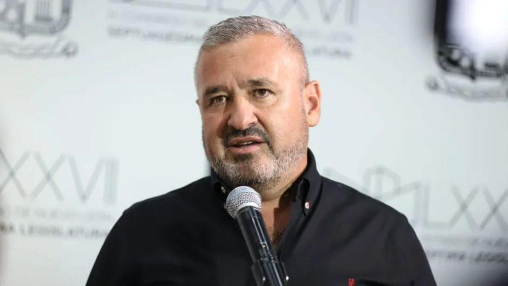 Man with gray beard speaking into a microphone at a podium or event wearing a black shirt   Informe Regio
