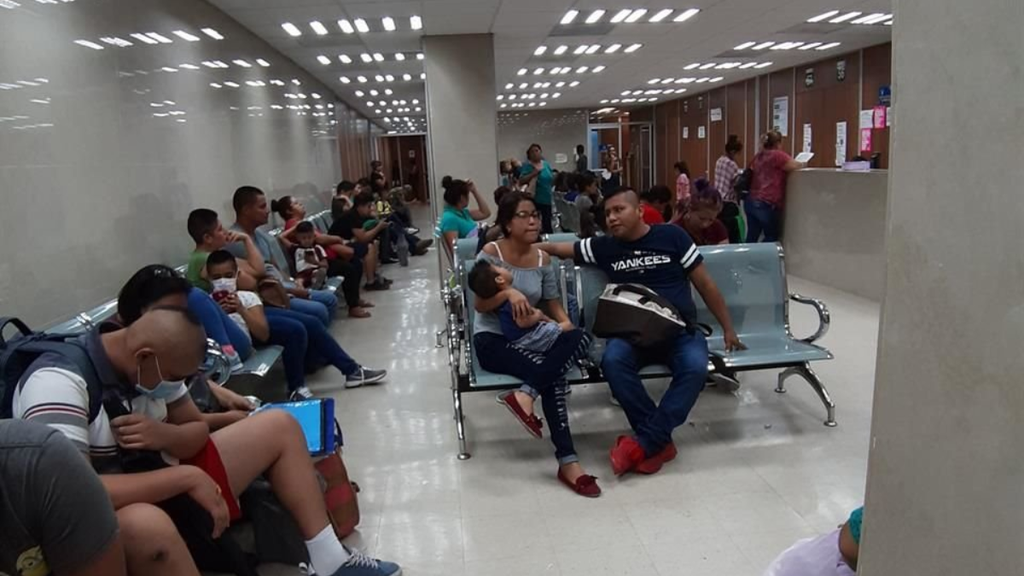 Crowded hospital waiting area with rows of seats filled by adults and children a man and woman holding a baby in the foreground and a reception desk in the back   Informe Regio