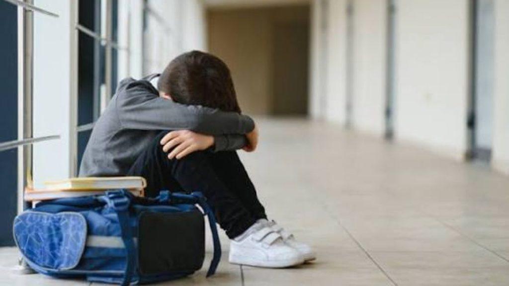 Student sitting on a tiled hallway floor with a backpack and books beside them head bowed and arms wrapped around their knees   Informe Regio